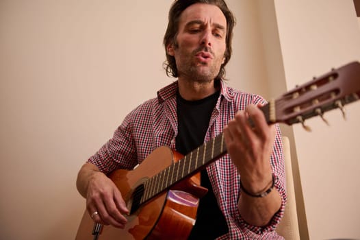 A man sits indoors, strumming a classical guitar while singing with focus and passion. The setting is relaxed, suggesting a personal or intimate practice session, capturing the beauty of music.