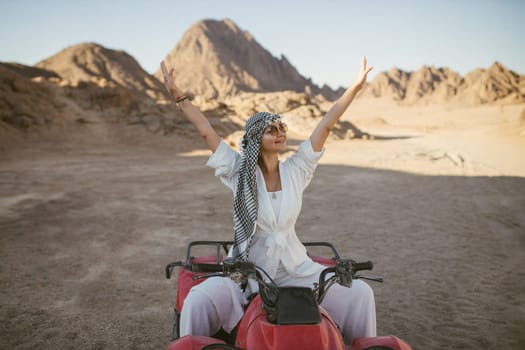 A rather attractive woman is going on a safari, driving an ATV in the desert. Beautiful young woman with a scarf on her head sits on a red four-wheeled ATV and rides towards adventure.
