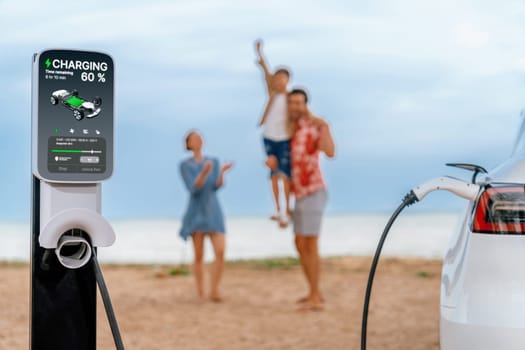 Alternative family vacation trip traveling by the beach with electric car recharging battery from EV charging station with blurred cheerful and happy family enjoying the seascape background. Perpetual