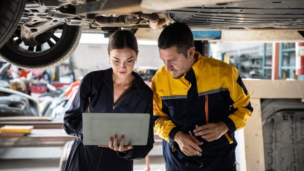 Two vehicle mechanic working together underneath lifted car, conduct car inspection with laptop. Automotive service technician in uniform carefully make diagnostic troubleshooting. Panorama Oxus