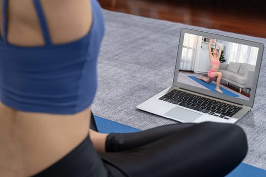 Asian woman in sportswear doing yoga exercise on fitness mat as her home workout training routine. Healthy body care lifestyle woman watching online yoga video on laptop. Vigorous