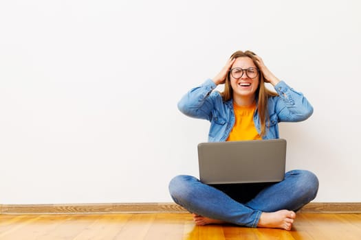 Stressed woman with laptop sitting on the floor against white wall holding her head making error in business project, missing deadline
