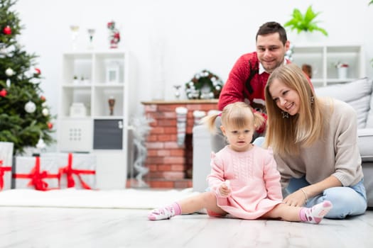 A family consisting of mom, dad and a little daughter pose for a photo in the room. The room is decorated in white and gray colors. A fireplace made of brick and a Christmas tree can be seen in the background.