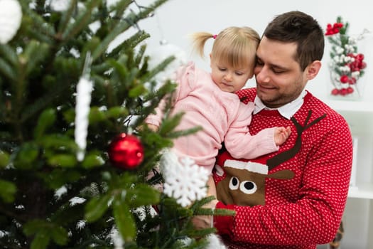 A man holds a little girl dressed in a pink dress in his arms. The man is wearing a holiday red sweater with an image of a reindeer. The man and the little girl are decorating a Christmas tree together.
