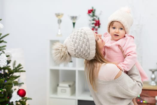 A mother carries her little daughter in her arms and smiles at her. The girl is wearing a pink dress and a cap. A fuzzy outline of a white bookcase can be seen in the background. A fragment of a Christmas tree can be seen next to the woman and her daughter.