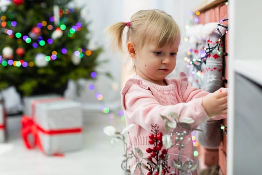 A tiny child watches Christmas tree lights hanging on a brick fireplace. The little girl is dressed in a pink dress. The child has light-colored hair. A decorated Christmas tree and presents can be seen in the background.
