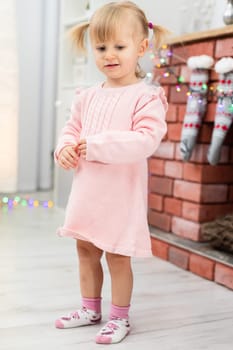 A tiny girl is standing in a room on a bright floor. The child is wearing a pink dress and colorful socks. The girl is standing by the fireplace, on which hang Christmas socks and colorful lights