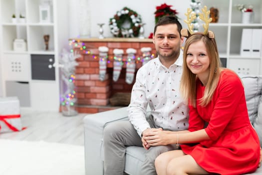 The girl and the boy are smiling and sitting on the gray couch. The couple embraces each other. The woman is wearing a red dress and the man is wearing a patterned shirt. In the background you can see a fireplace with Christmas socks hanging on it.
