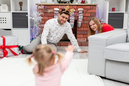 A couple in love sits on the floor by the fireplace. In the foreground is the blurred figure of a little girl running towards her parents. The room is decorated with Christmas ornaments