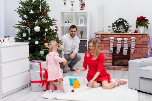 A blonde woman in a red dress sits on the carpet and plays with her little daughter. The little girl is standing on the carpet near her mother. A Christmas tree can be seen in the background. A man in a shirt is sitting by the Christmas tree.