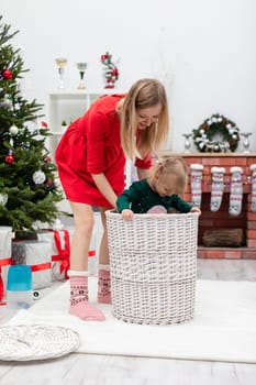A mother helps her little girl climb into a large wicker basket. The little girl plays with her mother. Mom and daughter play in a room full of Christmas decorations. The family spends the holidays together.
