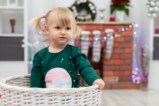 Close-up of a silhouette of a little girl sitting in a wicker basket. Blurred background. A brick fireplace can be seen in the background. Colorful Christmas lights are seen on the fireplace. The child is playing in the room.