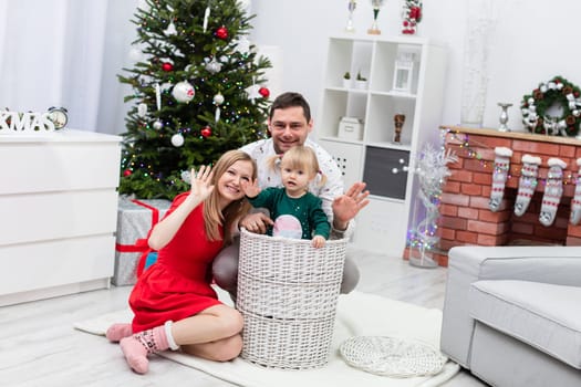 Mom and dad are sitting on the floor in a room decorated with Christmas ornaments. In front of the parents is a large wicker basket, in which stands a little girl. The child is dressed in a green sweatshirt. The parents are waving in front of them.