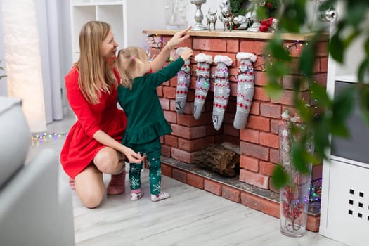 A woman dressed in a red dress crouches by the fireplace and embraces her daughter. The little girl stands by the fireplace and looks at the Christmas decorations. The brick fireplace is decorated with Christmas ornaments.
