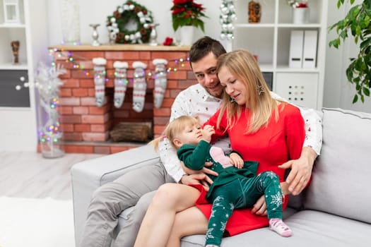Mom and dad hug their little daughter. The whole family is sitting on a gray couch. The little girl lies on her mother's lap. The woman is wearing a festive red dress. The family is celebrating the holidays.