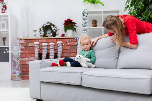 A woman in a red dress leans over a couch. A little boy is sitting on the couch. The mother is stroking her little son's cheek. Next to the boy lies a plush white toy.