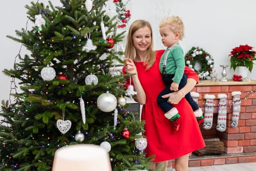 A young woman dressed in a red dress stands by a Christmas tree. The woman holds her little son in her arms. The boy is wearing a light green sweatshirt. The mother shows the child the ornaments on the Christmas tree.