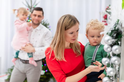 A mother holds a little boy in her arms. The woman shows the child the silver baubles on a Christmas wreath. A blurry figure can be seen in the background. A man holds a little girl in his arms and smiles.