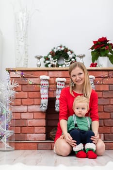 A smiling young mother holds her baby son on her lap. The family is spending time by the fireplace. The fireplace is decorated with Christmas lights and other holiday decorations.