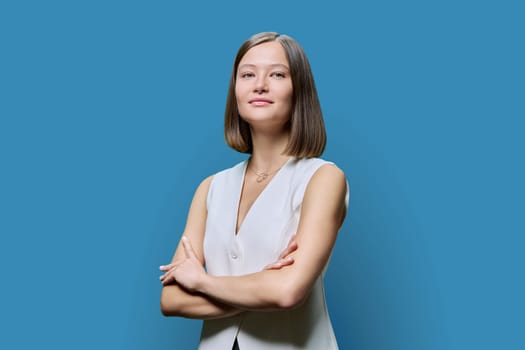 Portrait of young confident woman on blue studio background. Successful fashionable female with crossed arms looking at camera. Business, work, services, education, fashion beauty professions