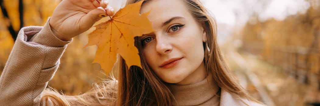Portrait of a woman with an autumn maple leaf. Railway, autumn leaves, a young long-haired woman in a light coat coat, close-up