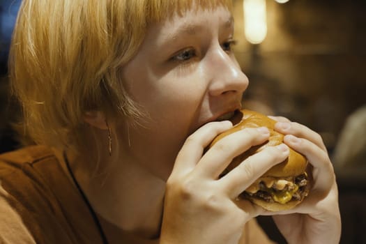A teenager girl eats a hamburger in a restaurant, enjoying a delicious juicy burger. Close-up.