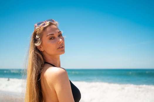 Blond woman on beach. Female with long hair and sunglasses on her head looking to camera while standing on sandy beach. Portrait of attractive girl in black swimwear with azure water and blue sky on backgrounds.