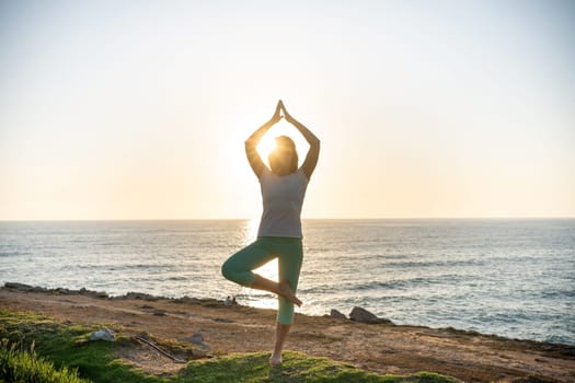 Senior woman practicing yoga on beach at sunset. Female doing yoga asana on coast with a sun setting on background. Silhouette of woman with slim body standing in yoga pose near ocean