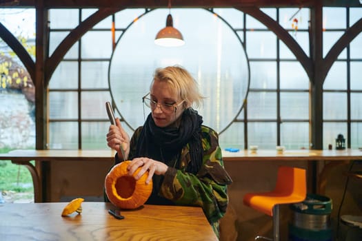 A modern blonde woman in military uniform is carving spooky pumpkins with a knife for Halloween night.