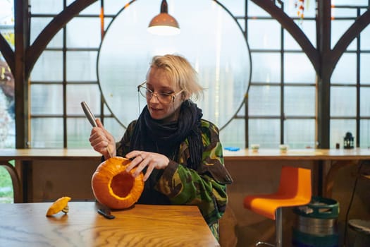 A modern blonde woman in military uniform is carving spooky pumpkins with a knife for Halloween night.