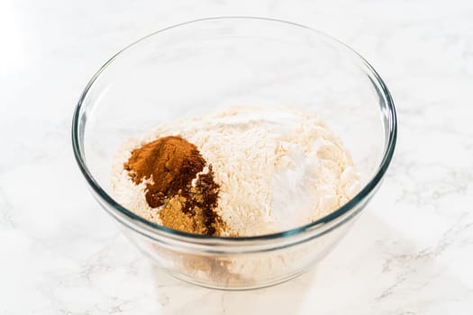 Close-up view of a large glass mixing bowl with flour and a measured amount of spices (including cinnamon and ginger) added on top, ready to be mixed for the Amber Gingerbread Cookie Dough.