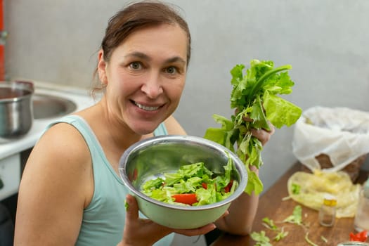 A woman shows a bowl of vegetables and herbs with lettuce leaves against the background of the kitchen.