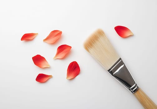 A makeup brush is placed beside scattered red rose petals on a clean white surface, creating a colorful composition.