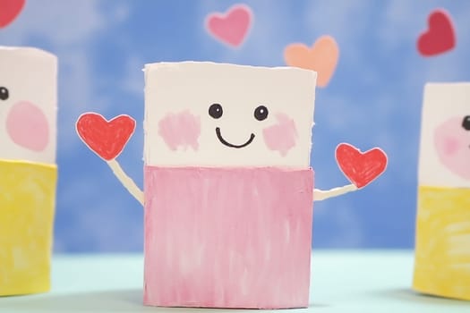 Cardboard figures with smiling faces and colorful outfits are joyfully holding red hearts against a cheerful blue background.