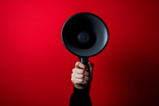 A person raises a black megaphone with a firm grip in front of a vibrant red background, ready to amplify their message.