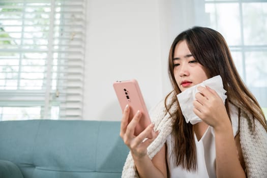 Asian young woman sick sitting down on sofa with cold blowing her nose and checking smart mobile phone to cell to doctor online, depressed sad sick female holding tissues and smartphone in living room