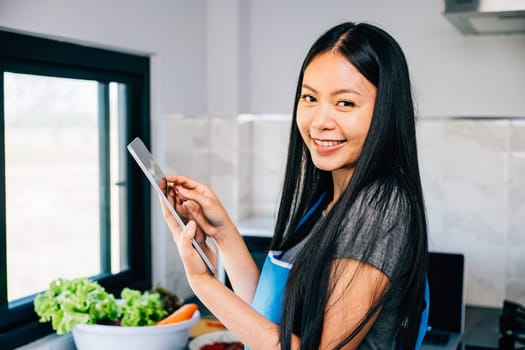 In a sun drenched kitchen a smiling Asian woman interacts with a tablet while cooking showcasing the joyful blend of technology and culinary artistry.