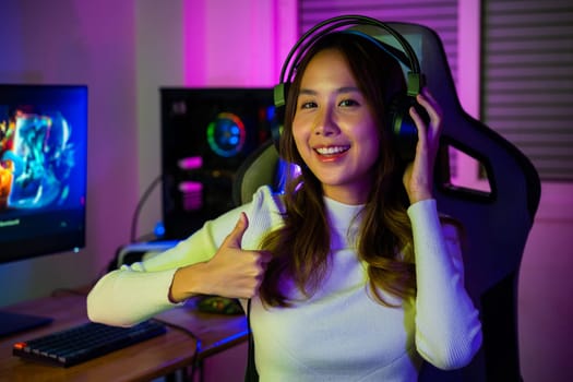 Gamer playing online game wear gaming headphones looking to camera expressing success with game giving thumbs up sign, Smiling woman live stream she play video game at home neon lights living room