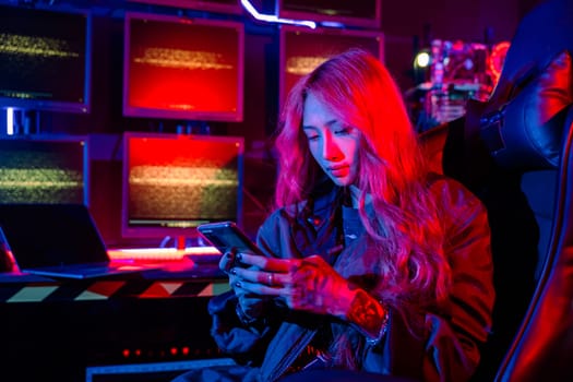 Asian woman in front of a computer monitor display using mobile phone, neon light, Hacker wear black jacket at system control room, Security guard working on multiple monitors together smartphone