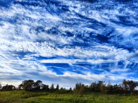 A bright sun shines over a green field with clouds streaking across a blue sky.
