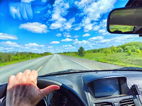 Car salon, windshield, hand of woman on steering wheel and landscape. View from seat of driver on nature with Road, tree and blue sky at sunny day
