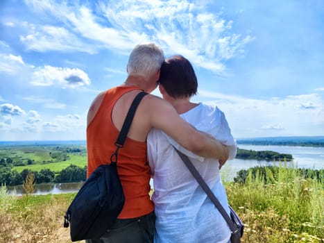 Two people embrace while admiring the lush countryside and river below under a bright blue sky with fluffy clouds.