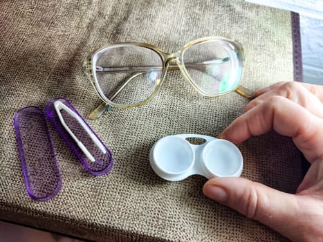 A hand holds a lens case while eyeglasses and accessories are placed on a textured surface, indicating preparation for lens use.
