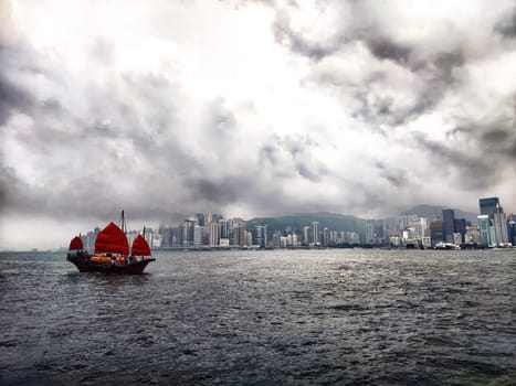 Hong Kong - April 05, 2024: A traditional ship with striking red sails navigates the waters of Victoria Bay, surrounded by impressive skyscrapers of Hong Kong under a moody sky filled with clouds.