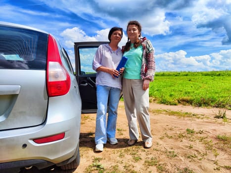 Women stand beside a silver car, enjoying the warm sun and expansive sky filled with clouds in a lush green field.