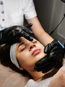 The beautician performs the procedure of microcurrents in black electric gloves to restore facial skin cells. Close-up, vertical