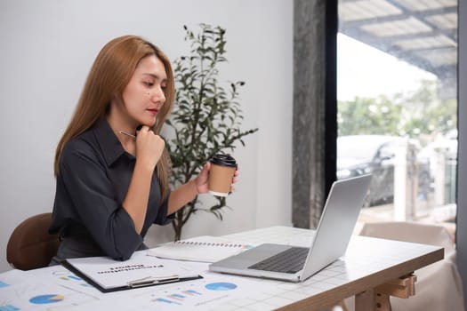 Young woman office worker analyzing data on a laptop, holding coffee, in a modern office environment.
