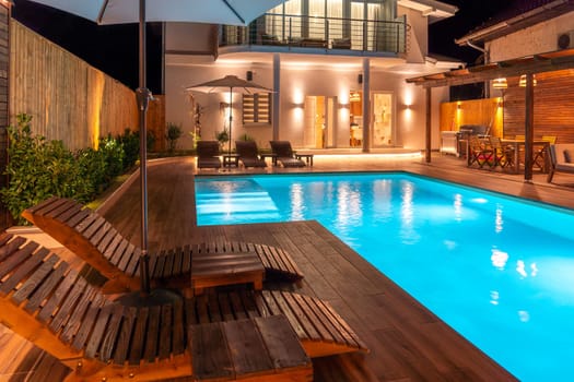 Luxury villa with illuminated swimming pool and wooden loungers at night. Concept of modern backyard relaxation