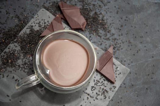 Glass cup of hot chocolate in heart shape on table surrounded by chocolate pieces and black chia seeds on grey textured background, Desert concept in color of the year 2025, mocha mousse,
