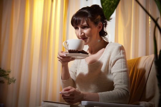 A middle-aged woman sits comfortably in her cozy apartment, sipping a warm beverage and enjoying a moment of relaxation during the evening.